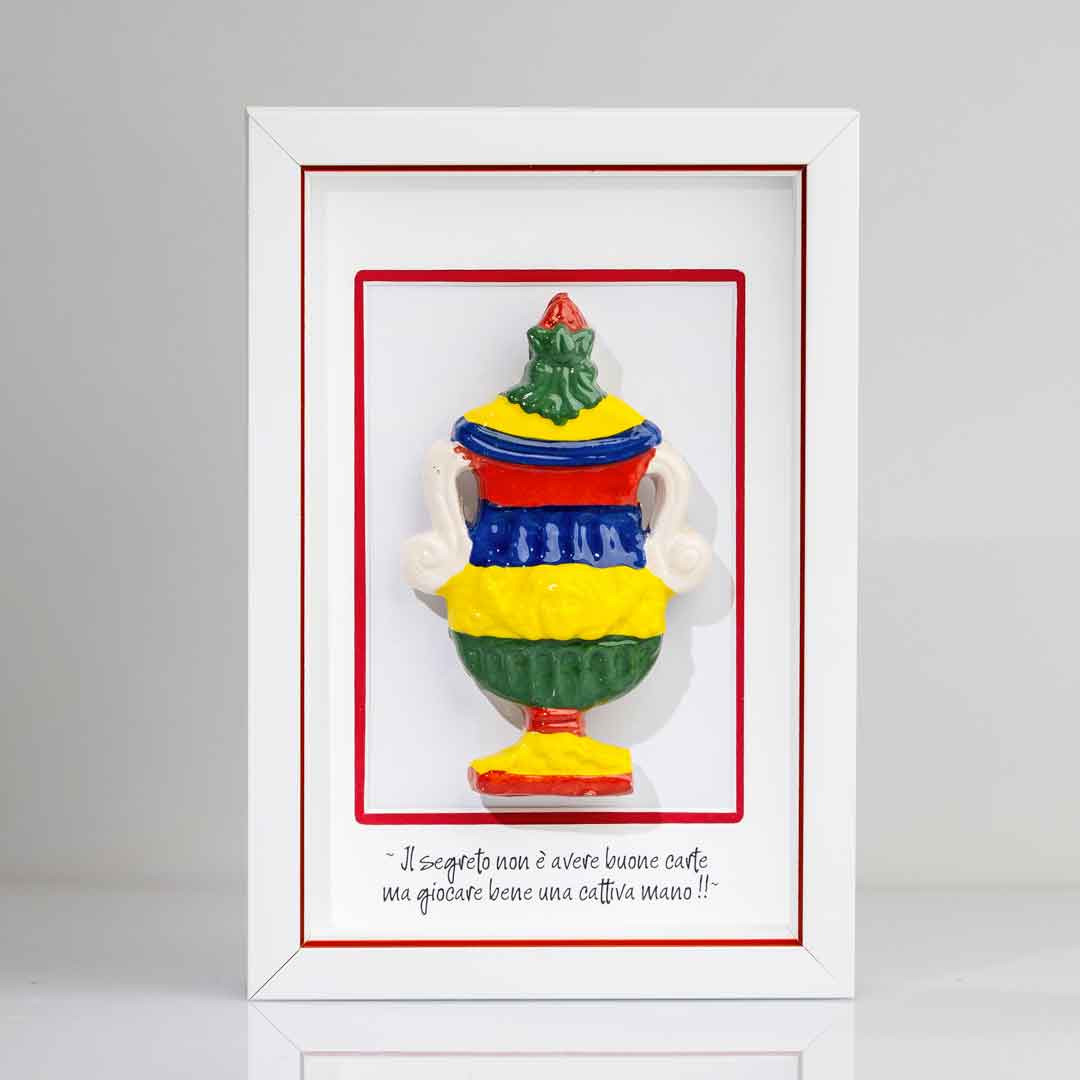 Quadro con cornice bianca contenente una riproduzione tridimensionale in ceramica colorata dell'Asso di Coppe delle carte napoletane. La coppa è decorata con bande orizzontali rosse, blu, gialle e verdi, e dettagli bianchi sui manici. In basso è riportata la scritta in corsivo: "Il segreto non è avere buone carte ma giocare bene una cattiva mano!!". cornice bianca con rigo rosso