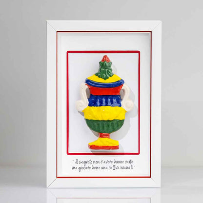 Quadro con cornice bianca contenente una riproduzione tridimensionale in ceramica colorata dell'Asso di Coppe delle carte napoletane. La coppa è decorata con bande orizzontali rosse, blu, gialle e verdi, e dettagli bianchi sui manici. In basso è riportata la scritta in corsivo: "Il segreto non è avere buone carte ma giocare bene una cattiva mano!!". cornice bianca con rigo rosso