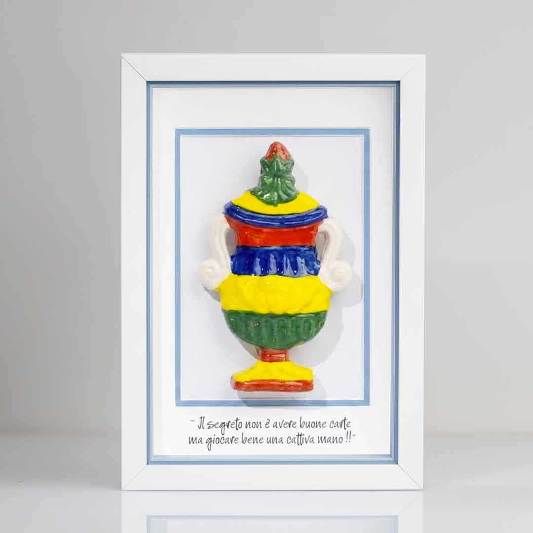 Quadro con cornice bianca contenente una riproduzione tridimensionale in ceramica colorata dell'Asso di Coppe delle carte napoletane. La coppa è decorata con bande orizzontali rosse, blu, gialle e verdi, e dettagli bianchi sui manici. In basso è riportata la scritta in corsivo: "Il segreto non è avere buone carte ma giocare bene una cattiva mano!!". Il bordo interno della cornice azzurro