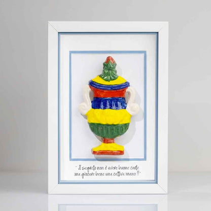 Quadro con cornice bianca contenente una riproduzione tridimensionale in ceramica colorata dell'Asso di Coppe delle carte napoletane. La coppa è decorata con bande orizzontali rosse, blu, gialle e verdi, e dettagli bianchi sui manici. In basso è riportata la scritta in corsivo: "Il segreto non è avere buone carte ma giocare bene una cattiva mano!!". Il bordo interno della cornice azzurro