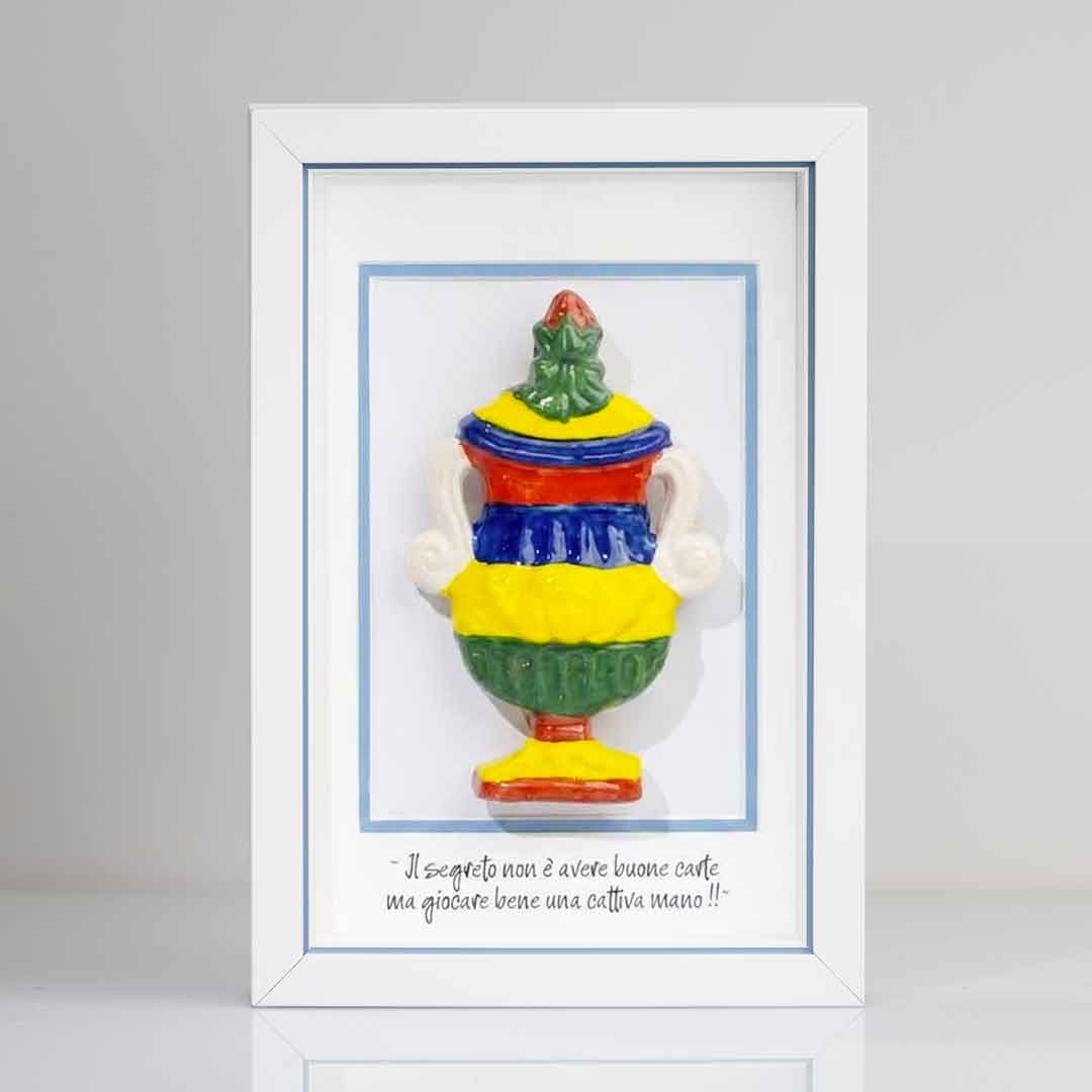 Riproduzione tridimensionale in ceramica colorata dell'Asso di Coppe delle carte napoletane. La coppa è decorata con bande orizzontali rosse, blu, gialle e verdi, e dettagli bianchi sui manici. In basso è riportata la scritta in corsivo: "Il segreto non è avere buone carte ma giocare bene una cattiva mano!!". cornice rigo azzurro
