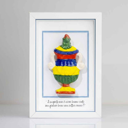 Riproduzione tridimensionale in ceramica colorata dell'Asso di Coppe delle carte napoletane. La coppa è decorata con bande orizzontali rosse, blu, gialle e verdi, e dettagli bianchi sui manici. In basso è riportata la scritta in corsivo: "Il segreto non è avere buone carte ma giocare bene una cattiva mano!!". cornice rigo azzurro