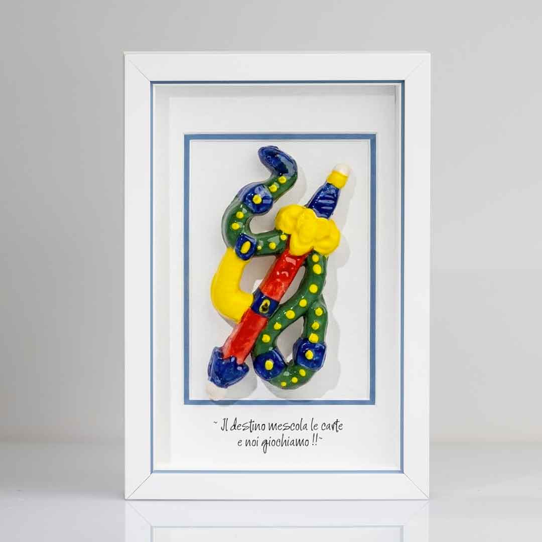 Quadro con cornice bianca contenente una riproduzione tridimensionale in ceramica dell'Asso di Spade delle carte napoletane. Raffigura una spada con impugnatura gialla e azzurra, avvolta da un elemento sinuoso verde a pois gialli. In basso è presente la scritta in corsivo: "Il destino mescola le carte e noi giochiamo!!". Il rigo interno della cornice e azzurro