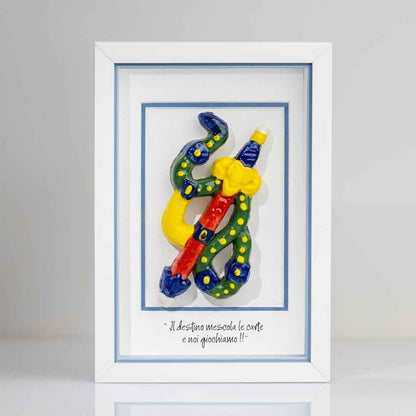 Quadro con cornice contenente una riproduzione tridimensionale in ceramica dell'Asso di Spade delle carte napoletane. Raffigura una spada con impugnatura gialla e azzurra, avvolta da un elemento sinuoso verde a pois gialli. In basso è presente la scritta in corsivo: "Il destino mescola le carte e noi giochiamo!!". Il bordo interno della cornice è azzurro