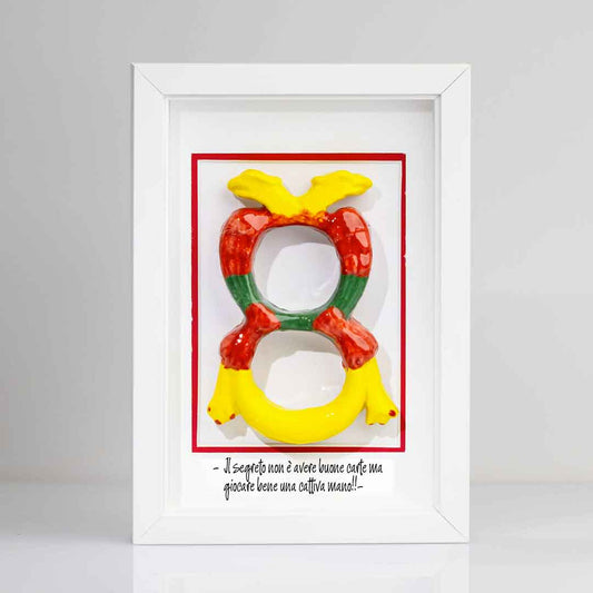 Quadro con contenente una riproduzione tridimensionale in ceramica dell'Asso di Denari delle carte napoletane, raffigurato come una grande moneta o sole stilizzato con decorazioni geometriche e antropomorfe nei toni del giallo, rosso e verde. In basso è riportata la scritta in corsivo: "Il segreto non è avere buone carte ma giocare bene una cattiva mano!!". Cornice tutta bianca