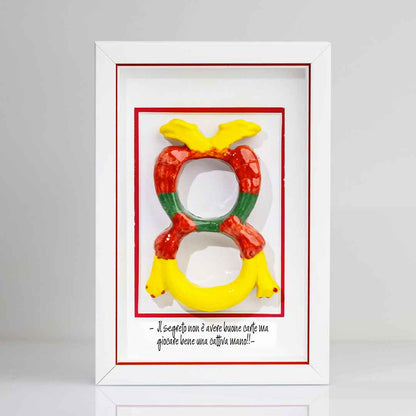 Quadro contenente una riproduzione tridimensionale in ceramica dell'Asso di Denari delle carte napoletane, raffigurato come una grande moneta o sole stilizzato con decorazioni geometriche e antropomorfe nei toni del giallo, rosso e verde. In basso è riportata la scritta in corsivo: "Il segreto non è avere buone carte ma giocare bene una cattiva mano!!". Il rigo interno della cornice è rosso