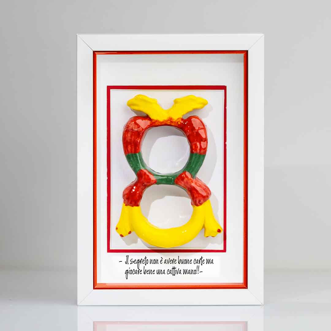 Quadro contenente una riproduzione tridimensionale in ceramica dell'Asso di Denari delle carte napoletane, raffigurato come una grande moneta o sole stilizzato con decorazioni geometriche e antropomorfe nei toni del giallo, rosso e verde. In basso è riportata la scritta in corsivo: "Il segreto non è avere buone carte ma giocare bene una cattiva mano!!". Il bordo interno della cornice è rosso