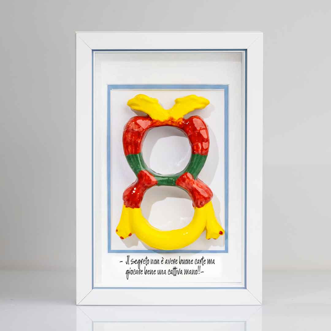 Quadro con cornice contenente una riproduzione tridimensionale in ceramica dell'Asso di Denari delle carte napoletane, raffigurato come una grande moneta o sole stilizzato con decorazioni geometriche e antropomorfe nei toni del giallo, rosso e verde. In basso è riportata la scritta in corsivo: "Il segreto non è avere buone carte ma giocare bene una cattiva mano!!". Il bordo interno della cornice è azzurro