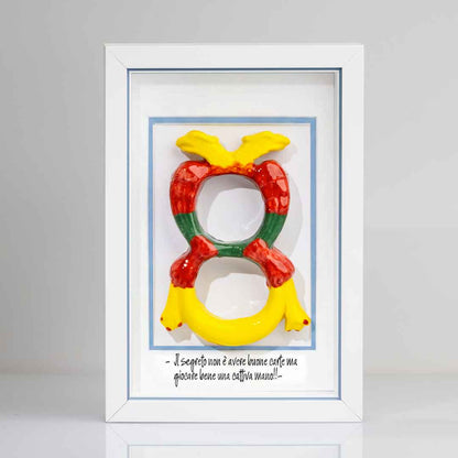 Quadro con cornice contenente una riproduzione tridimensionale in ceramica dell'Asso di Denari delle carte napoletane, raffigurato come una grande moneta o sole stilizzato con decorazioni geometriche e antropomorfe nei toni del giallo, rosso e verde. In basso è riportata la scritta in corsivo: "Il segreto non è avere buone carte ma giocare bene una cattiva mano!!". Il bordo interno della cornice è azzurro