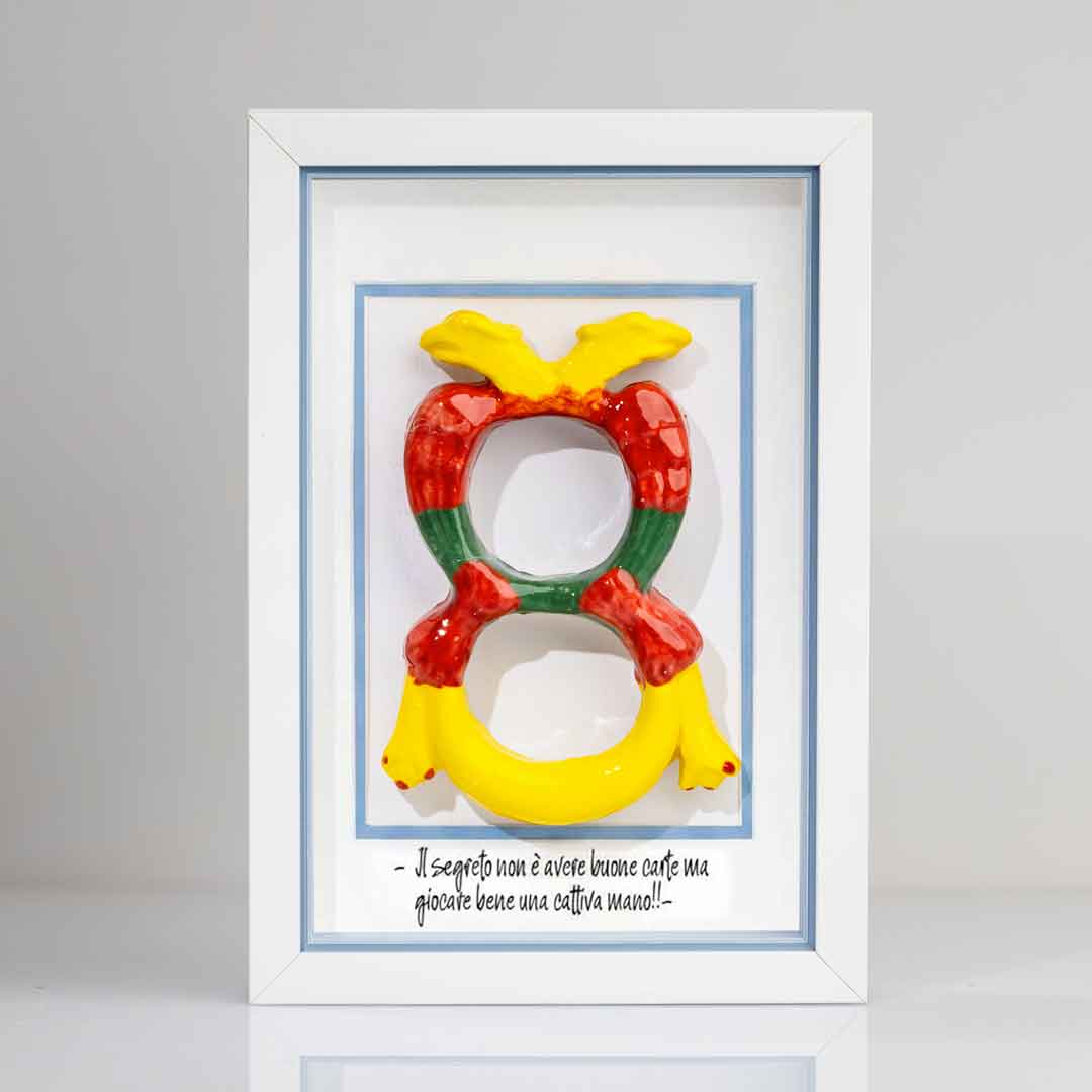Quadro con cornice contenente una riproduzione tridimensionale in ceramica dell'Asso di Denari delle carte napoletane, raffigurato come una grande moneta o sole stilizzato con decorazioni geometriche e antropomorfe nei toni del giallo, rosso e verde. In basso è riportata la scritta in corsivo: "Il segreto non è avere buone carte ma giocare bene una cattiva mano!!". Il bordo interno della cornice è azzurro