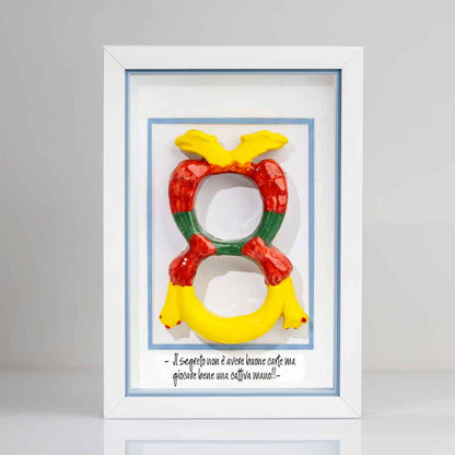 Quadro con cornice contenente una riproduzione tridimensionale in ceramica dell'Asso di Denari delle carte napoletane, raffigurato come una grande moneta o sole stilizzato con decorazioni geometriche e antropomorfe nei toni del giallo, rosso e verde. In basso è riportata la scritta in corsivo: "Il segreto non è avere buone carte ma giocare bene una cattiva mano!!". Il bordo interno della cornice è azzurro